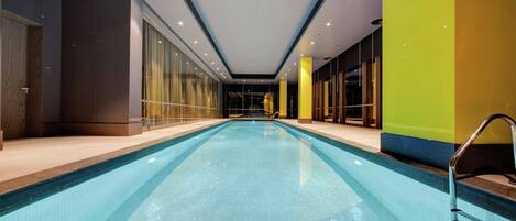 A modern indoor swimming pool with clear blue water, surrounded by glass walls and a tiled floor.
