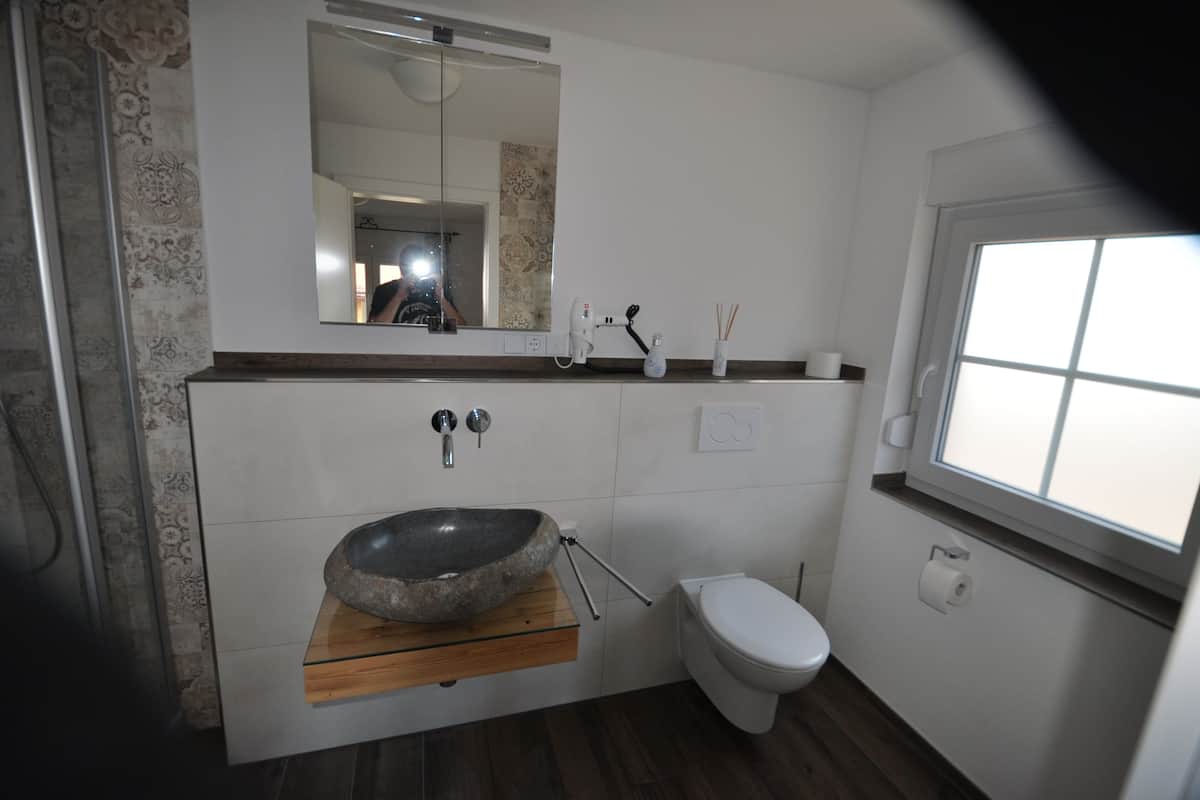 Een badkamer met een stenen wastafel, toilet, spiegel en raam.