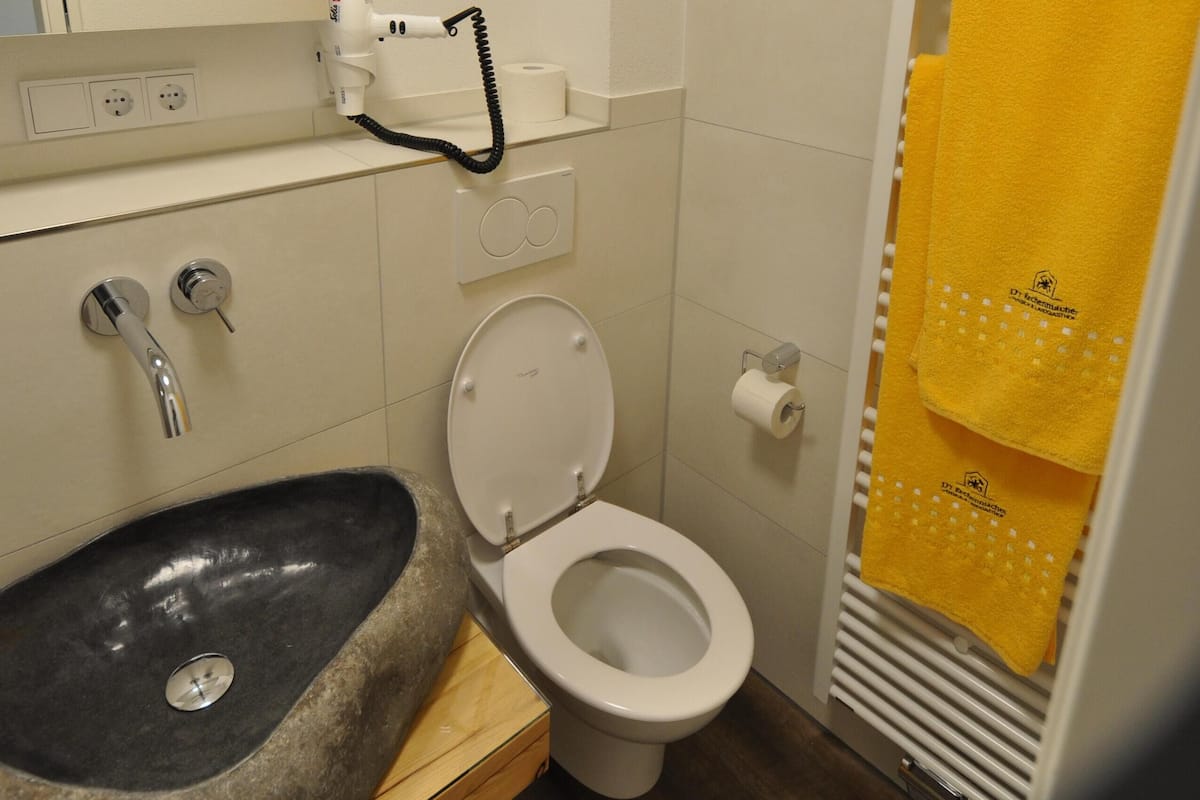 Een badkamer met een stenen wastafel, een wit toilet, een spiegel en een gele handdoek die aan een rek hangt.
