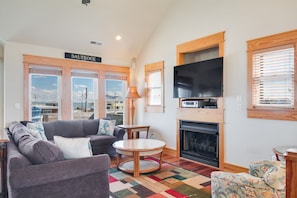 A living room with a sofa, a round table, a flat-screen TV, a fireplace, and large windows with blinds.