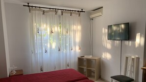 Ein Zimmer mit zwei Betten, einem Fernseher, einem Sessel, einem kleinen Tisch und einem Fenster mit Vorhängen.