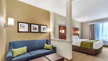 A hotel room with a blue sofa, a bed with a green blanket, a brown ottoman, and framed pictures on the wall.