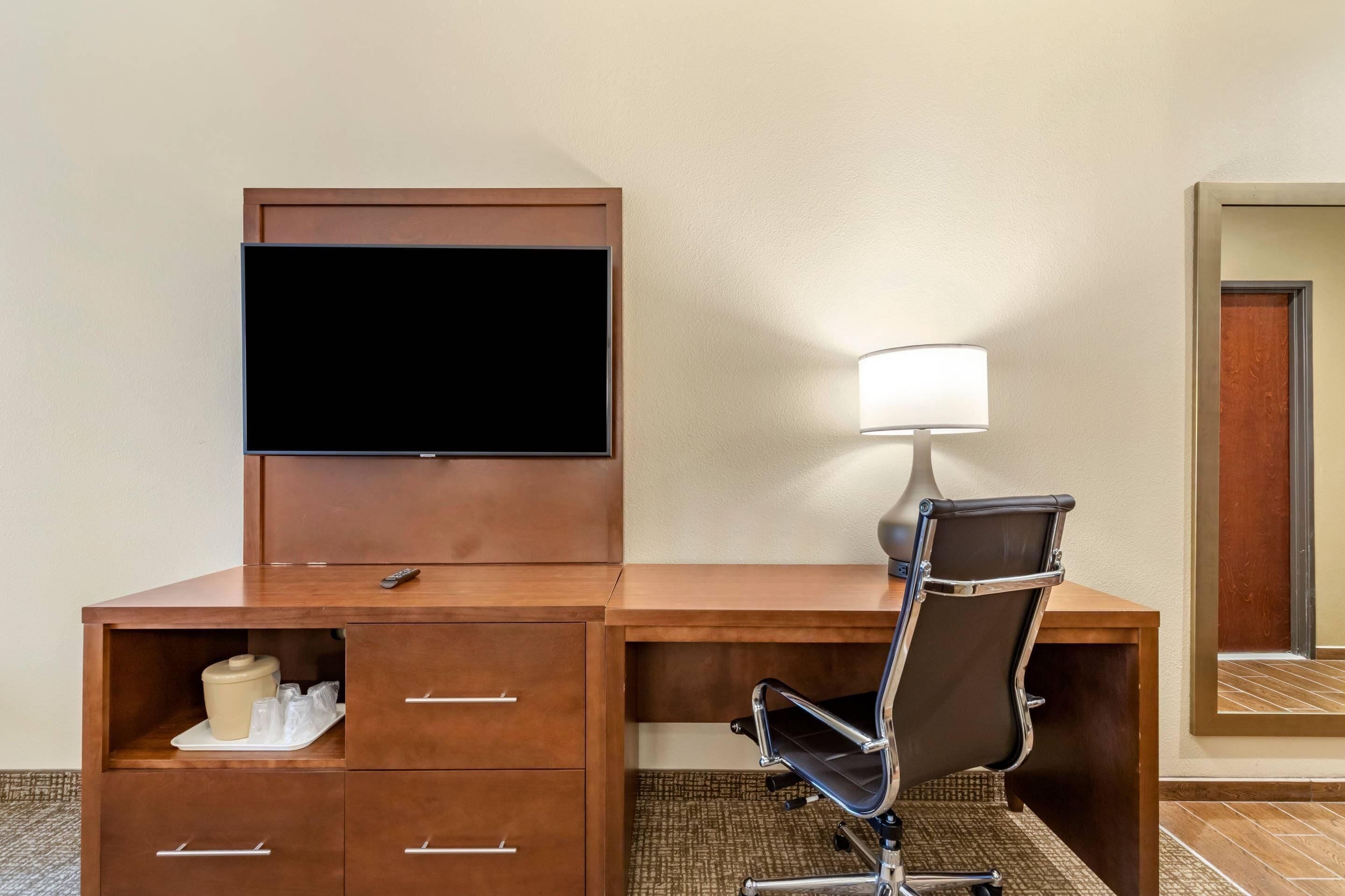 Ein Hotelzimmer mit Schreibtisch aus Holz, einem Stuhl, einem Fernseher, einer Lampe und einer Minibar.
