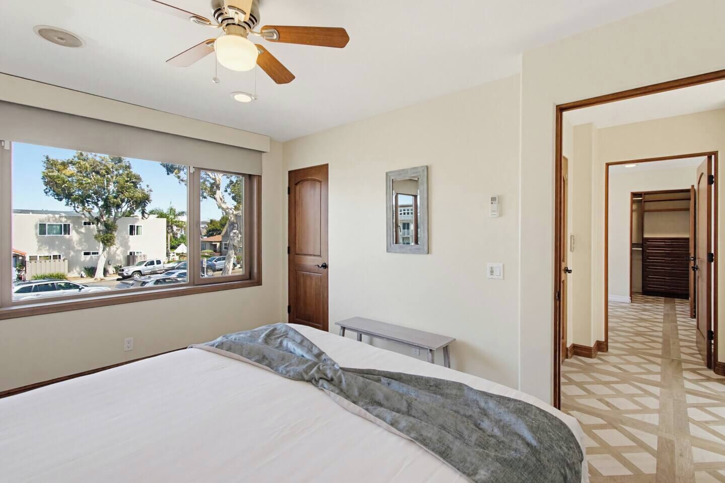 A bedroom with a bed, a ceiling fan, a window with a view of parked cars and trees, and a hallway leading to a closet.