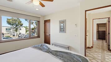 A bedroom with a bed, a ceiling fan, a window with a view of parked cars and trees, and a hallway leading to a closet.