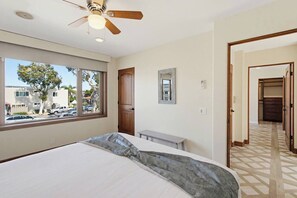 A bedroom with a bed, a ceiling fan, a window with a view of parked cars and trees, and a hallway leading to a closet.