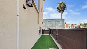 A narrow alleyway with artificial green turf, a brown fence, and a building with windows and a door. - New Photos! Luxury Steps to Beach w/hot tub garage (San Diego)