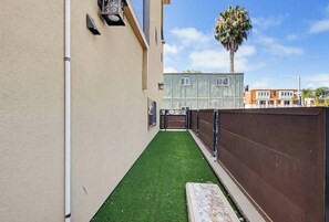 A narrow alleyway with artificial green turf, a brown fence, and a building with windows and a door.