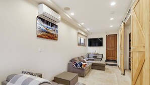 A living room with a sofa, a television, and a wall-mounted air conditioner. - New Photos! Luxury Steps to Beach w/hot tub garage (San Diego)