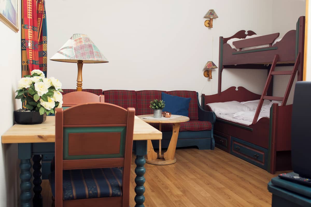 A cozy room with a wooden desk, a chair, a small table with a lamp, a sofa, a bunk bed, and a potted plant.