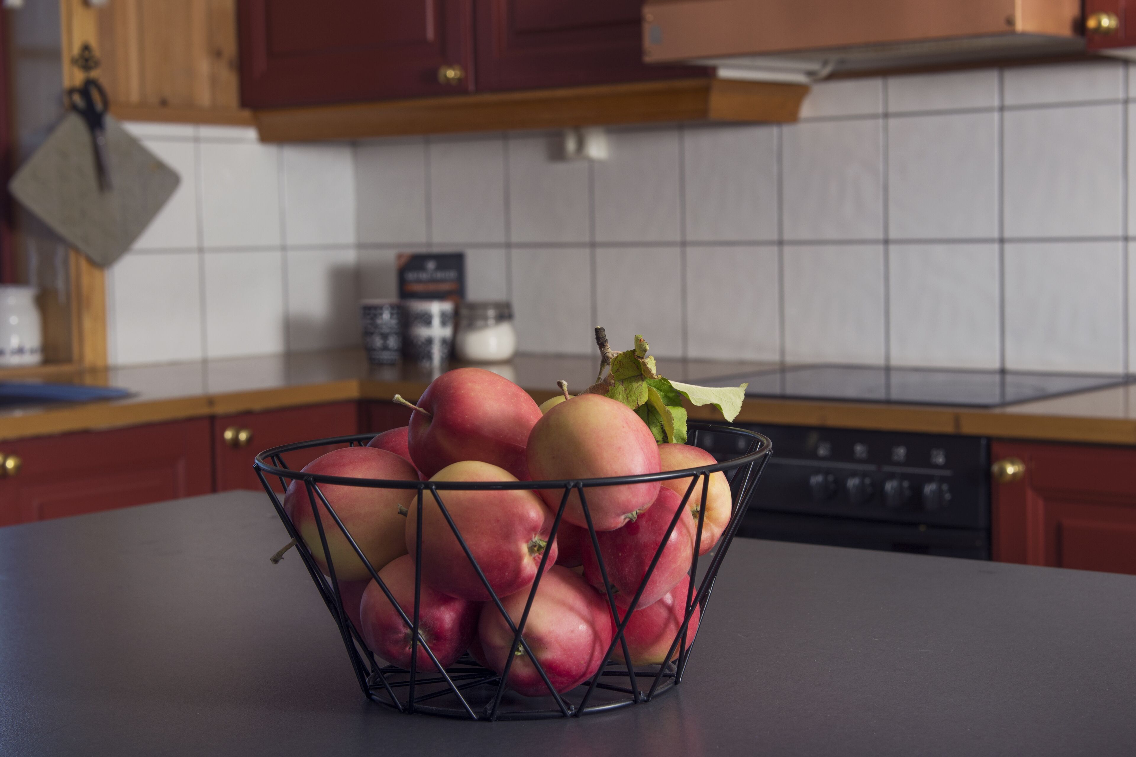 A kitchen with wooden cabinets, a tiled backsplash, and a countertop with a wire fruit basket containing apples.
