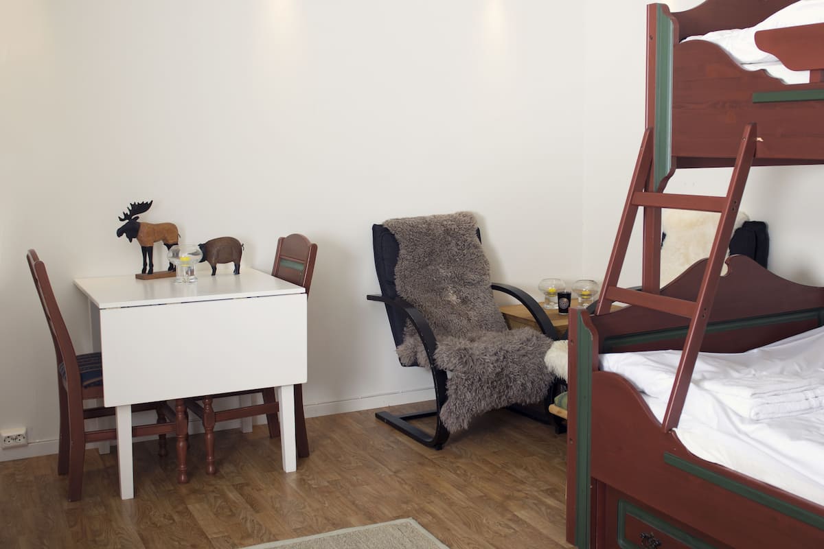 A room with a bunk bed, a small table with wooden chairs, a fluffy chair, and a wall-mounted shelf with decorative items.
