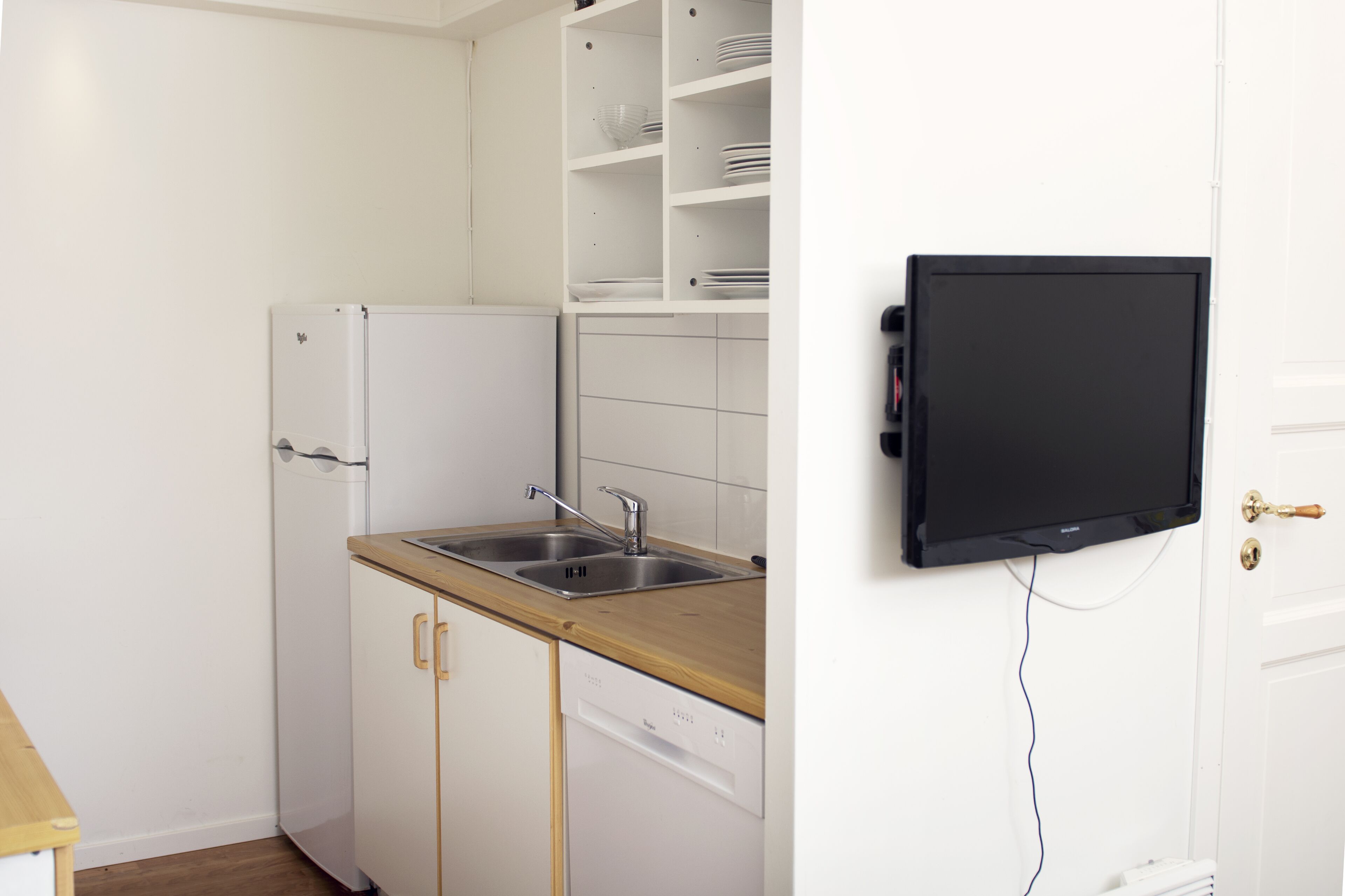 A compact kitchen with white cabinetry, a wooden countertop, a stainless steel sink, and a black television mounted on the wall.
