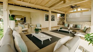 Ein großzügiger Wohnbereich mit hohen Decken, Holzbalken und einer Steinwand. Es gibt mehrere Sitzgruppen mit Sofas und Sesseln sowie einen Couchtisch in der Mitte.