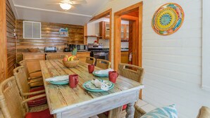 Un coin repas accueillant avec une table en bois, des chaises en osier et un saladier rempli de fruits. On y trouve également un ventilateur de plafond et une décoration murale colorée.