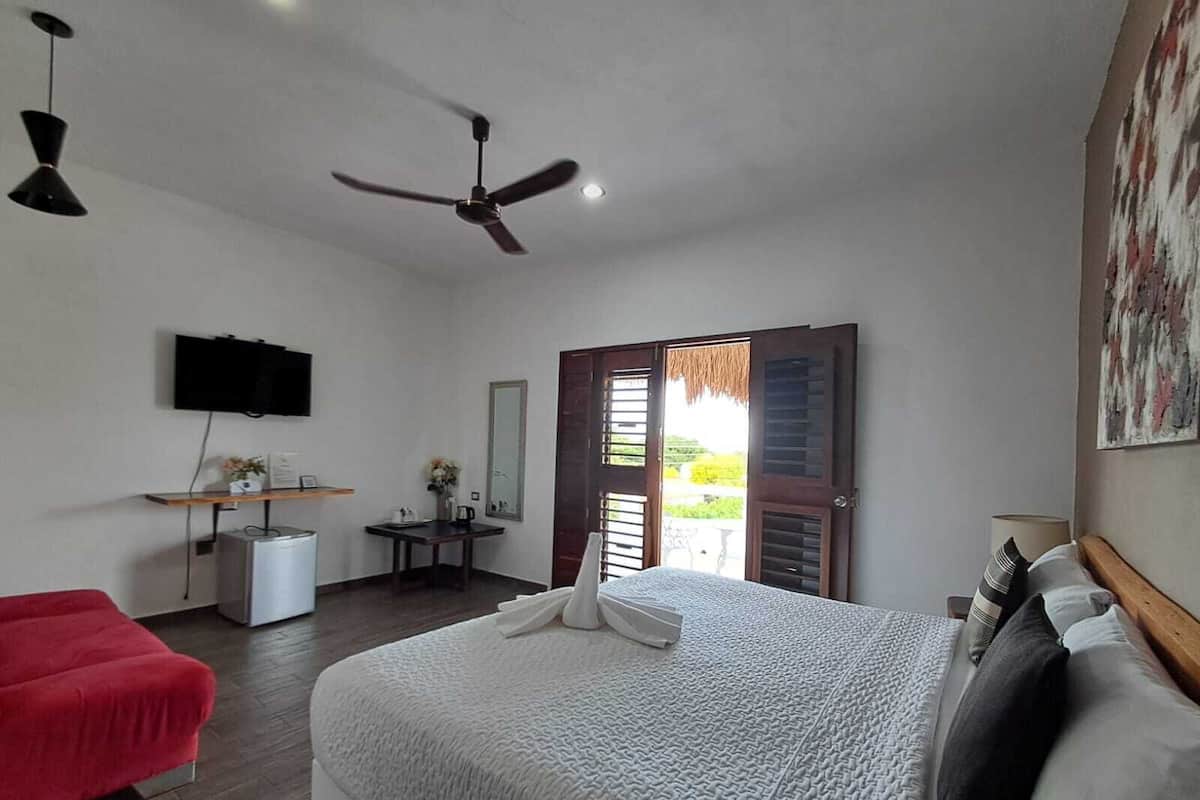 A bedroom with a bed, a red chair, a desk with a mirror, a television, and a ceiling fan.