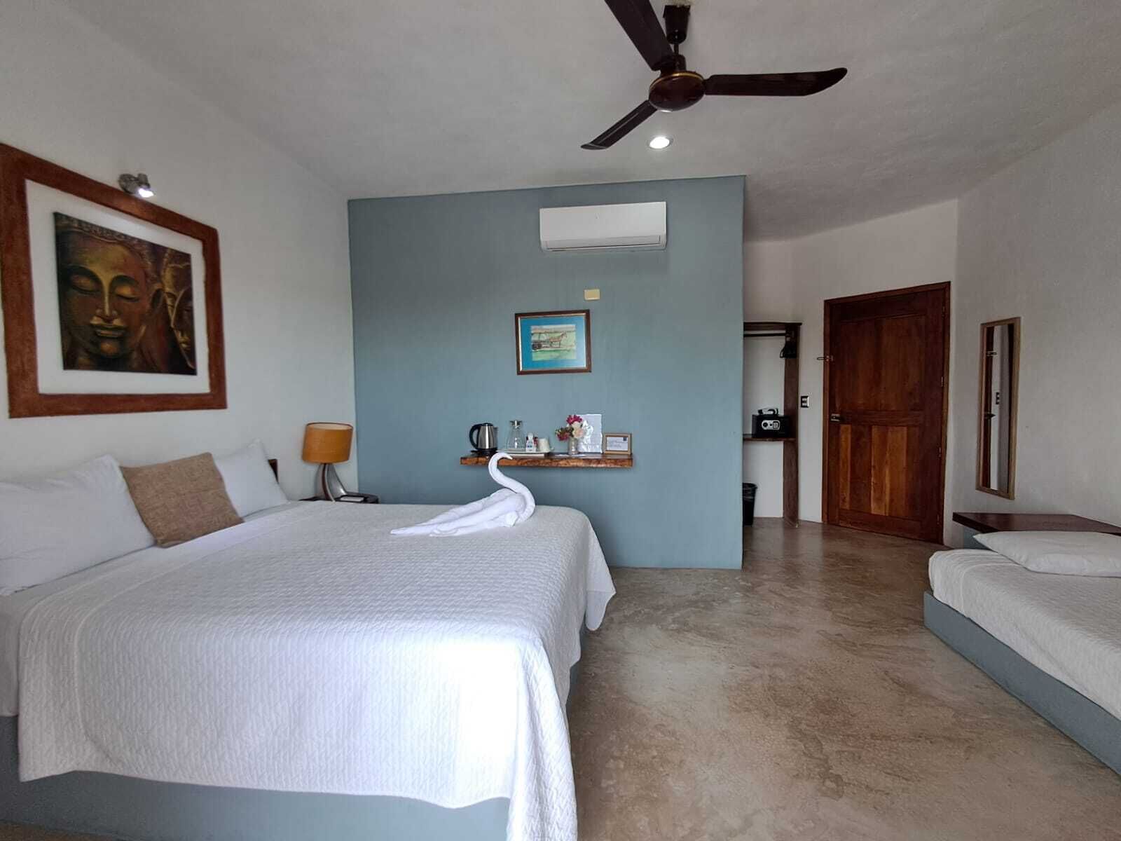 Habitación de hotel con dos camas, un ventilador de techo, una estatua de Buda enmarcada, un estante pequeño con una cafetera y una puerta que da a otra habitación.