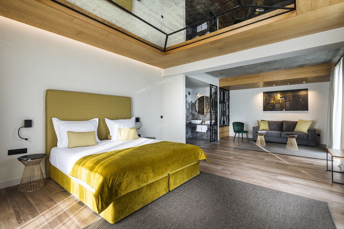 A modern bedroom with a large bed, a wooden ceiling, and a view of a living area with a sofa and chairs.