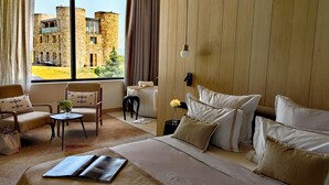 A hotel room with a large bed, a sitting area with a table and chairs, a view of a stone building outside, and a wooden wall panel. - Maison & Chateau Copsa (Karlovo)