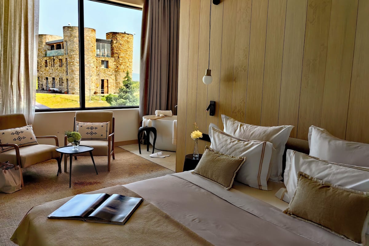 A hotel room with a large bed, a sitting area with a table and chairs, a view of a stone building outside, and a wooden wall panel.