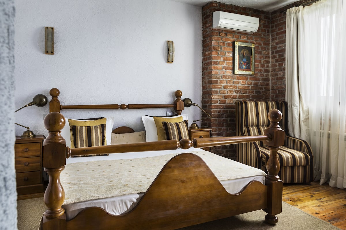 A traditional wooden bed with a headboard, a nightstand, a striped armchair, and a brick wall with a picture.