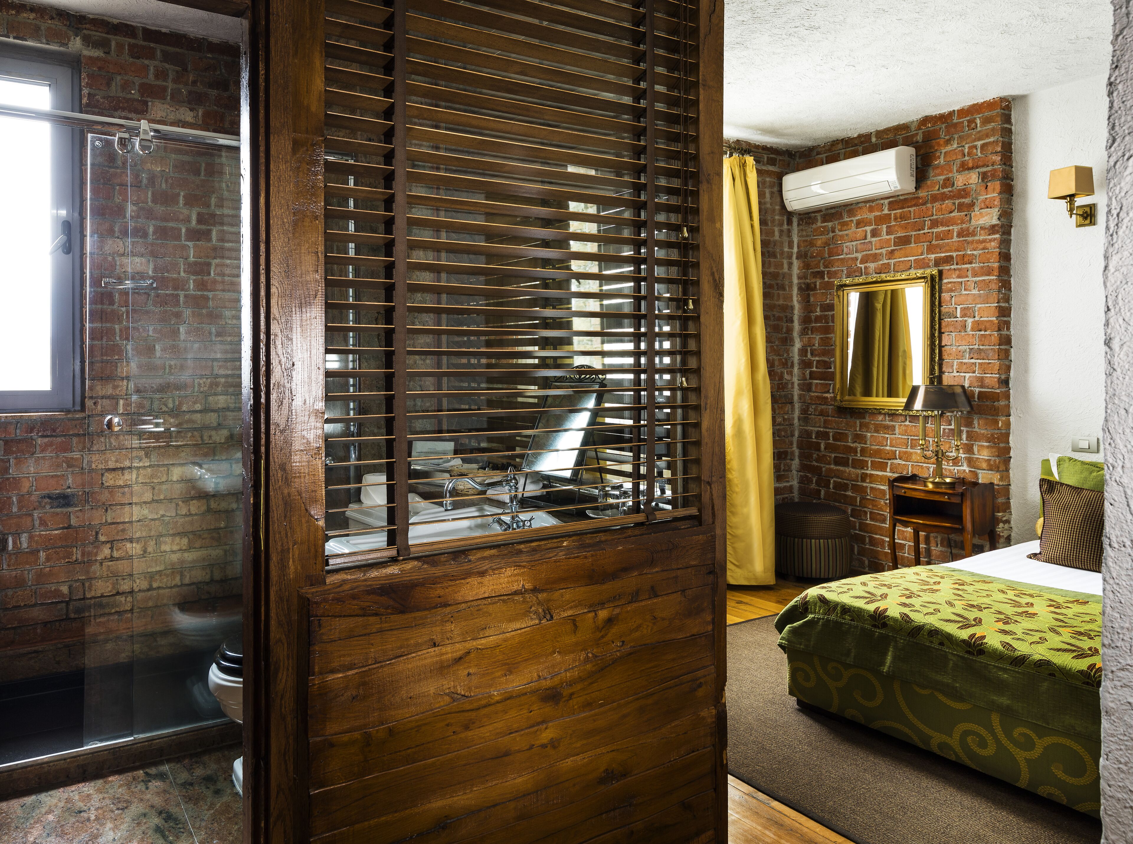A room with a wooden door, a bathroom with a shower, a bed with a green patterned bedspread, a small wooden nightstand, and a wall-mounted mirror.
