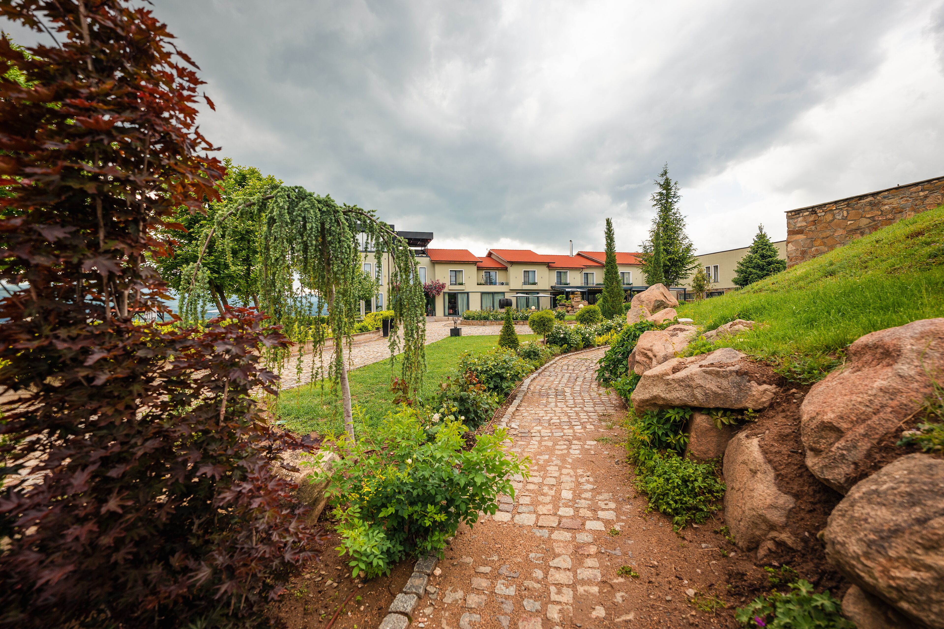 A pathway leading to a building with a red roof, surrounded by greenery and rocks.