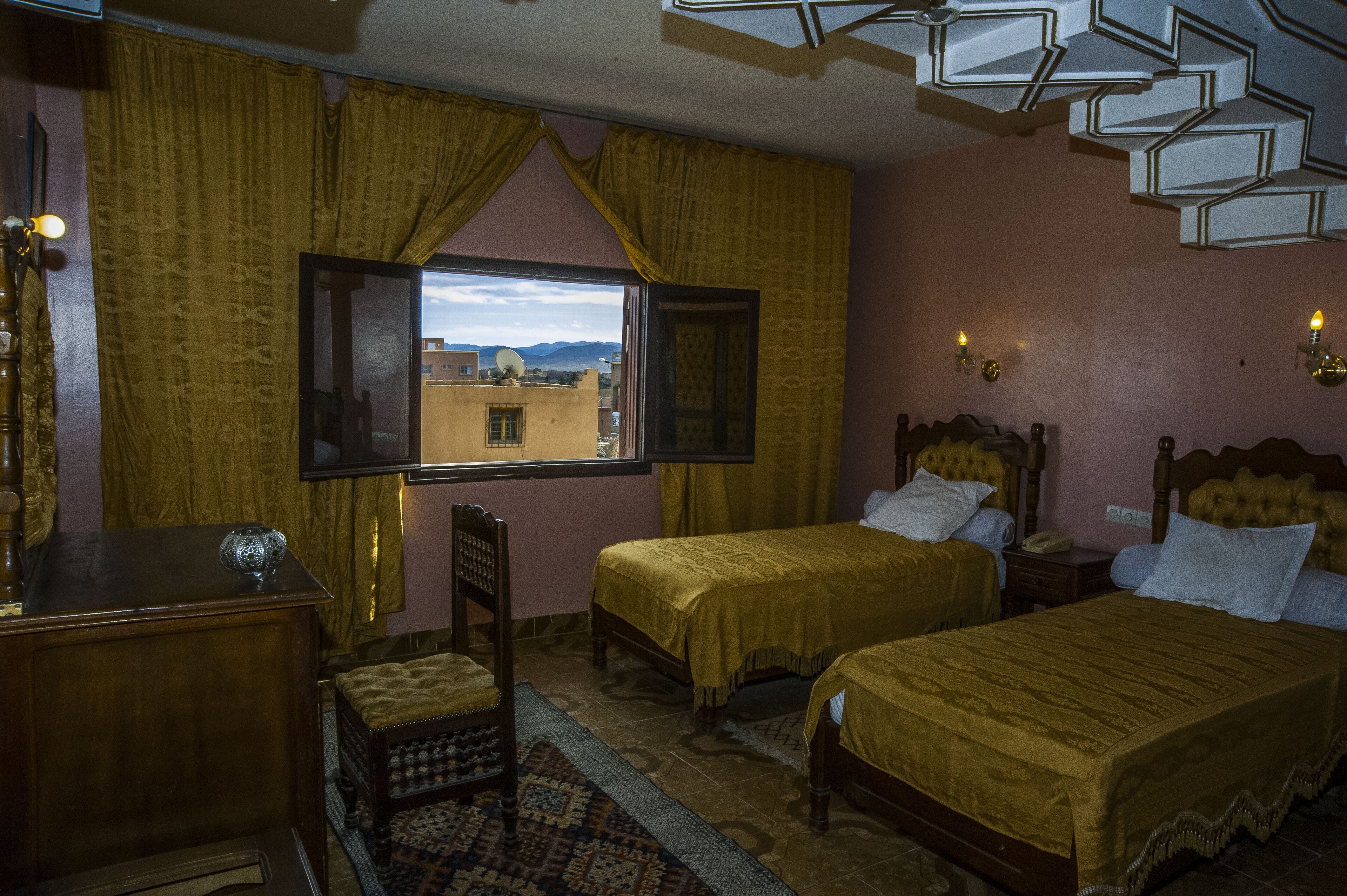 A room with two beds, a wooden desk, a chair, a window with curtains, and a patterned rug.