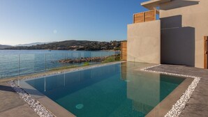 Een modern buitenzwembad met helderblauw water, omringd door een terras met kiezelstenen en uitzicht op een kalm meer met heuvels in de verte.