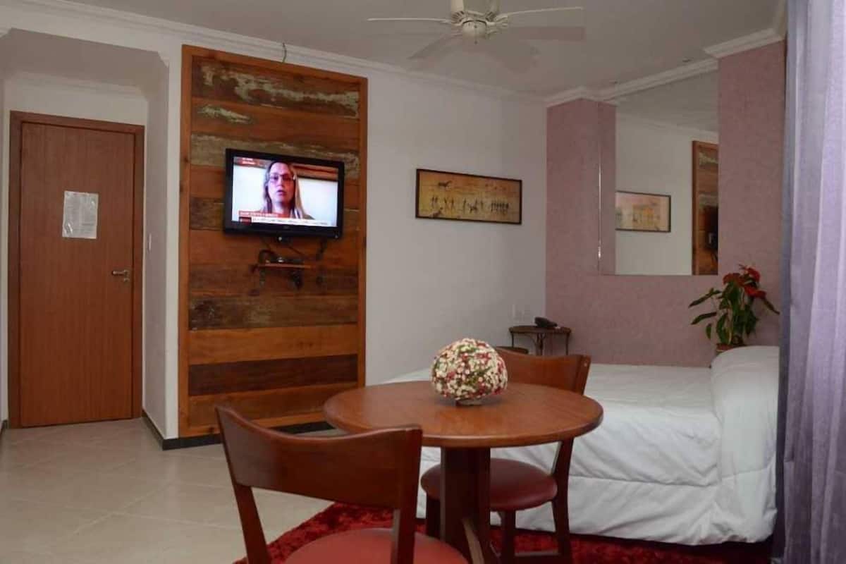 A hotel room with a wooden accent wall, a television, a round table with chairs, a bed, and a small table with a plant.