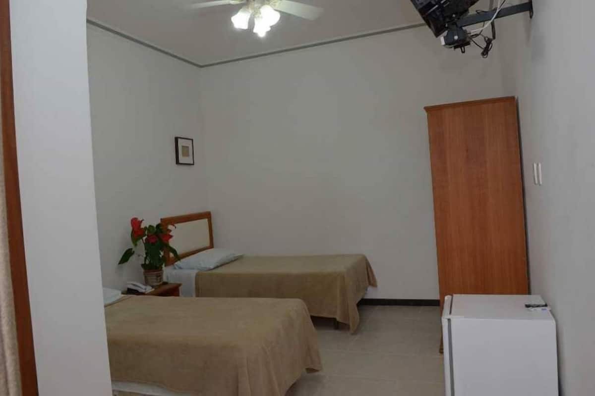 A room with two single beds, a wooden wardrobe, a ceiling fan, and a white refrigerator.