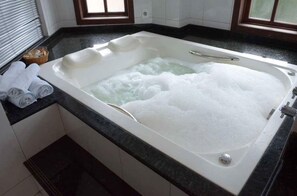 Una tina blanca grande llena de burbujas, un perchero con toallas y una ventana con persianas.