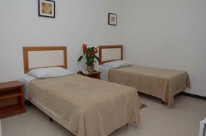 Una habitación con dos camas individuales, cada una con cabecero de madera y ropa de cama color beige. Hay una mesita de noche de madera con un teléfono y una planta en maceta con flores rojas.