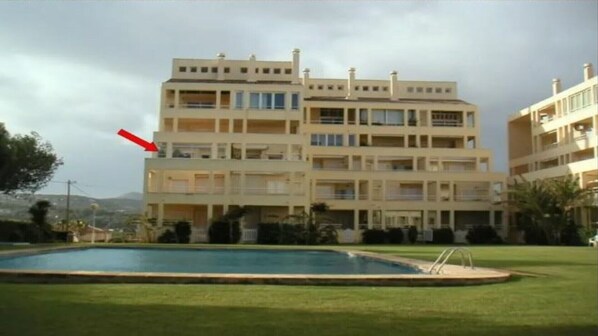 Residential building with a flat roof and balconies on each floor, set amidst lush greenery and a hilly landscape.