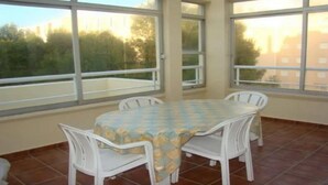 A room with a tiled floor, a round table with a checkered tablecloth, and four white chairs. Large windows provide a view of trees and buildings outside. - apartment sea view large terrace (Xàbia)
