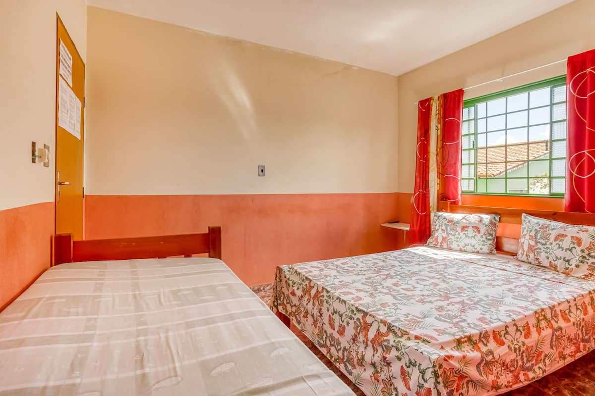 A room with two beds, one with a floral bedspread and the other with a plain one. There is a window with red curtains and a view of buildings outside.