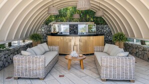 Un área de estar exterior techada con muebles de mimbre, una mesa redonda central y una barra con fondo de piedra.