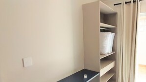 A closet with shelves and hanging space, a power outlet, and a window with curtains. - Residencial Miragem Bombinhas Duo (Bombinhas)