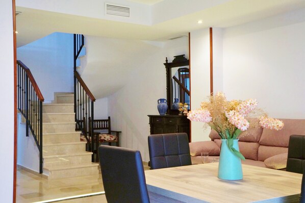 Un coin repas avec une table en bois, des chaises et un vase de fleurs. On aperçoit un escalier au fond, qui mène à une autre pièce.