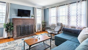 Ein Wohnzimmer mit Kamin aus Ziegelsteinen, einem flachbildfernseher darüber, einem blauen Sofa, einem Holztisch und einem Teppich mit Muster.