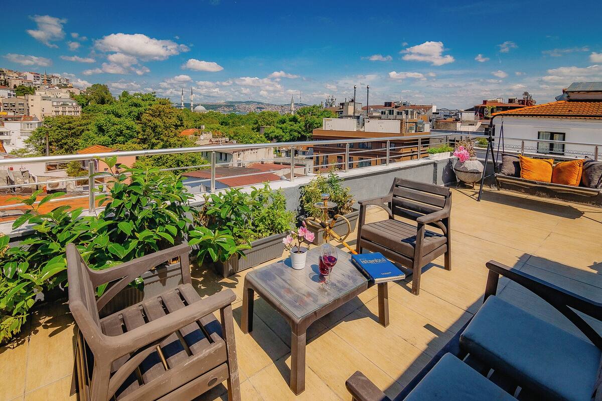 A rooftop terrace with outdoor furniture, a table with a book and a glass of wine, and a view of the cityscape.