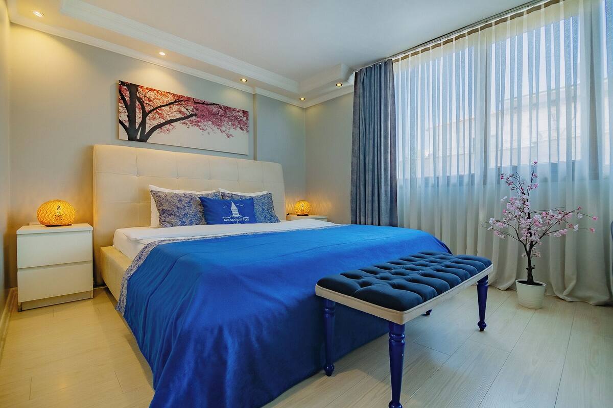 A modern bedroom with a large bed, a bedside table with a lamp, a bench, and a potted plant.