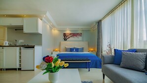 Ein modernes Hotelzimmer mit Küchenzeile, einem Bett mit blauer Bettwäsche, einem grauen Sofa und einer Vase mit Blumen auf einem weißen Tisch.