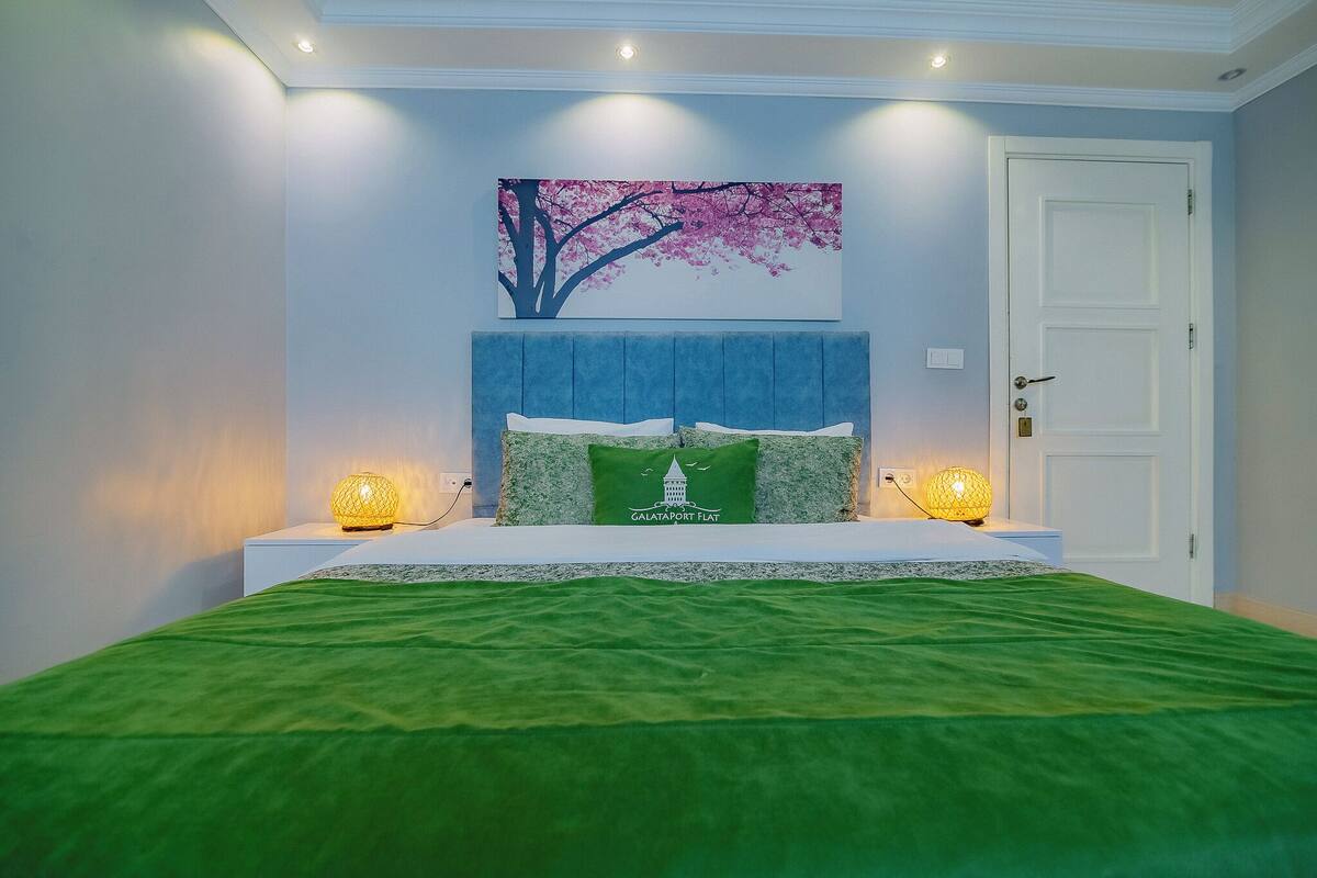 A bedroom with a bed, two bedside lamps, a wall-mounted painting, and a door.