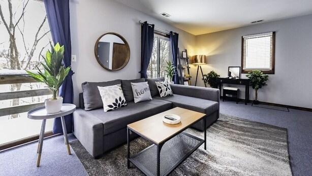 A modern living room with a grey sofa, a coffee table, and a small side table with a plant. There is a large mirror on the wall, a lamp, and a window with curtains. - Secluded 2BR Loft - Minutes from Downtown AA! (Ann Arbor)