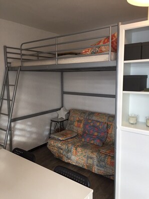 Ein Stockbett mit einer Leiter, ein kleiner Tisch mit einer Lampe, ein farbenfrohes Sofa und ein weißes Schränkchen mit Regalen.