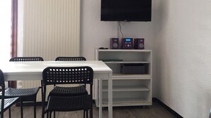 A room with a white table, two black chairs, a white shelf with speakers, and a mounted television. - C - Refurbished accommodation / sleeps 4 (Lans-en-Vercors)