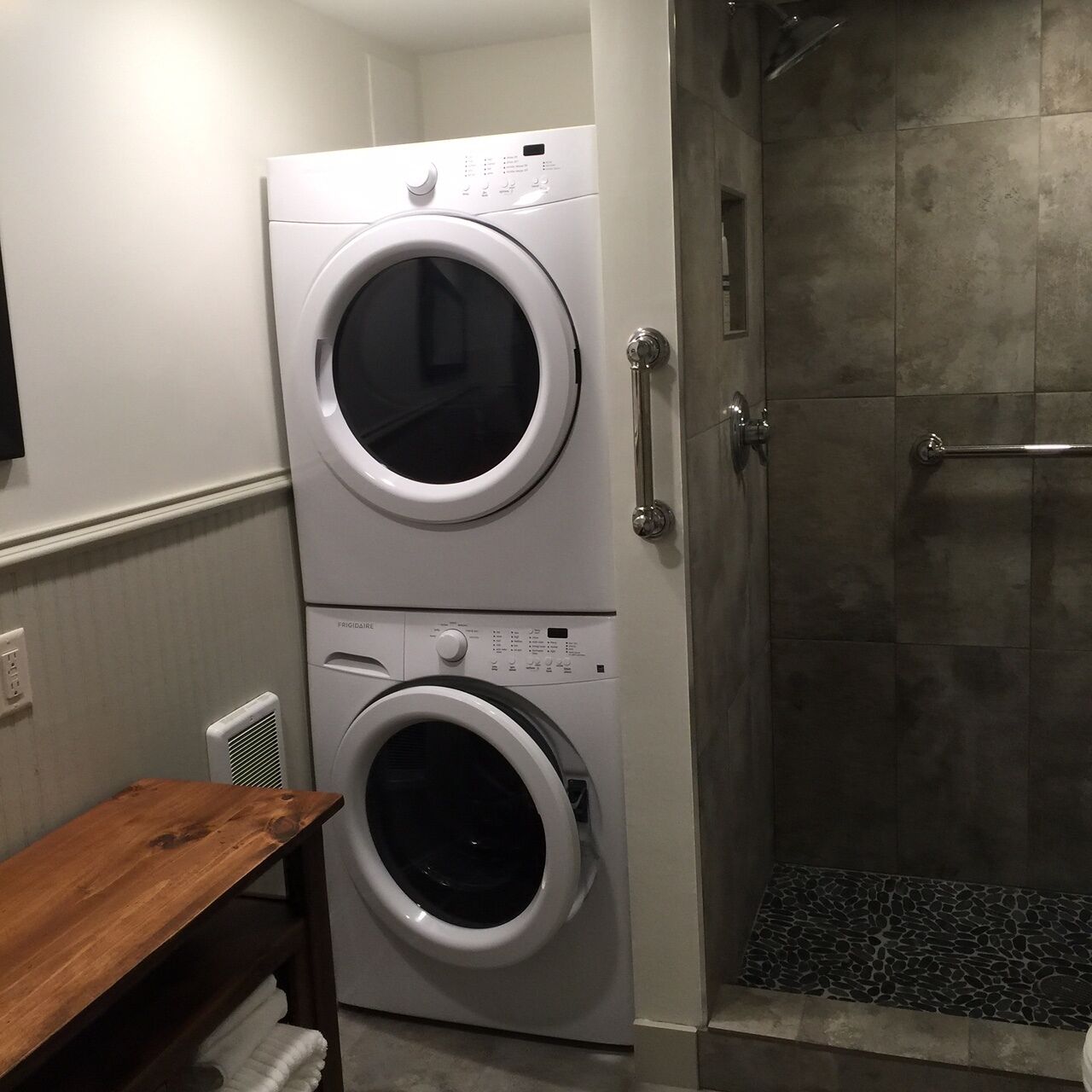 A laundry room with two stacked front-loading washing machines, a wooden bench, and a shower area.