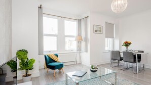A modern living room with a glass coffee table, a blue armchair, a dining area with a table and chairs, and large windows with curtains.