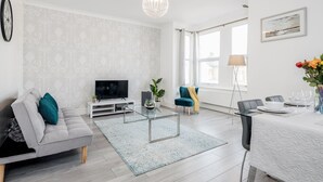 A modern living room with a sofa, TV, glass coffee table, and dining area with chairs and table set for two.
